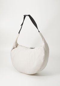 Bolsa acolchada con forma redondeada y semiovalada. Fabricada en tejido beige claro, cuenta con un cierre con cremallera y una correa negra ajustable.