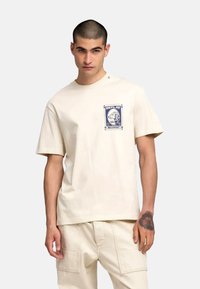 Camiseta beige de manga corta con un estampado gráfico en el lado izquierdo del pecho, que presenta un diseño azul y la palabra "REPLAY". Confeccionada con material de algodón suave.
