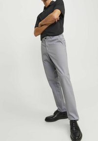 Persona con braccia incrociate, indossando una polo nera, pantaloni grigio chiaro e scarpe di pelle nere, in piedi contro uno sfondo chiaro e uniforme.