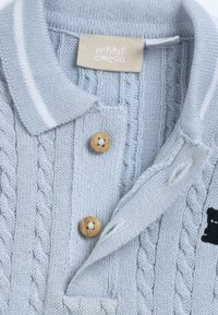 Cardigan pour bébé en tricot bleu clair avec un motif de torsades, boutons en bois, col avec rayures blanches et une petite broderie d'ours.