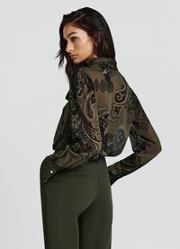 Blusa trasparente nera e verde oliva con motivo paisley. Maniche lunghe, polsini con bottoni e una silhouette fluida. Indossata con pantaloni verde oliva.