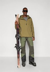 Équipement de ski comprenant un pull vert avec une fermeture éclair, une couche de base grise, un pantalon cargo vert à plusieurs tons et des chaussures de ski noires tenant des skis.