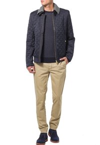 Marinblå quiltad jacka med grå krage, bärs över en marinblå tröja med prickar. Beiga chinos och marinblå skor fulländar outfiten.