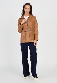 Brun læderbæltejakke med en rund krave, to frontlommer og en afslappet pasform, parret med mørk denim bukser med vide ben.