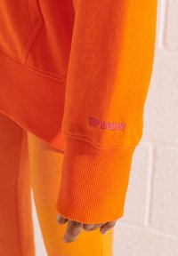 Sudadera naranja con puños acanalados, que presenta "SUPERDRY" bordado en púrpura. Textura suave y corte relajado. Pantalones naranjas a juego visibles.