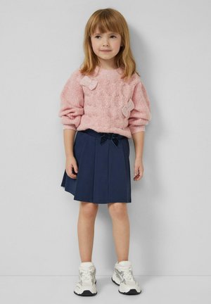 Jasnoróżowy sweter dziergany z teksturowanym wzorem i dwoma kokardkami, w połączeniu z granatową plisowaną spódnicą i białymi trampkami.