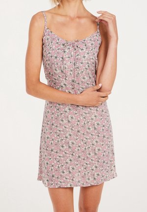 Kobieta ubrana w różową, kwiecistą sukienkę bez rękawów z cienkimi ramiączkami i małą kokardką na wysokości klatki piersiowej, stojąca na białym tle.