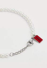 Pärlhalsband med jämna vita pärlor, silverdetaljer och en röd tagg märkt 'HUGO', med en hummerlåsning och kedjeutvidgning.