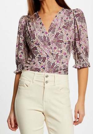 Femme portant un chemisier cache-cœur à motif floral avec des manches trois-quarts bouffantes et un pantalon crème taille haute à doubles boutons.