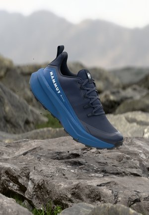 Tumši zils Mammut pārgājienu apavu pāris ar zilu zoli, piekārts virs klinšainas takas, fonā redzamas miglainas kalnu ainavas.