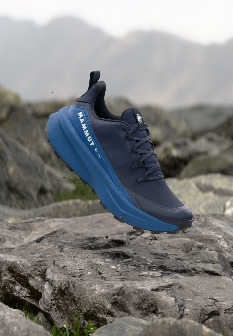 Σκούρο μπλε παπούτσι πεζοπορίας Mammut με μπλε σόλα κρεμασμένο πάνω από πετρώδες έδαφος με θολό φόντο ορεινών περιοχών.