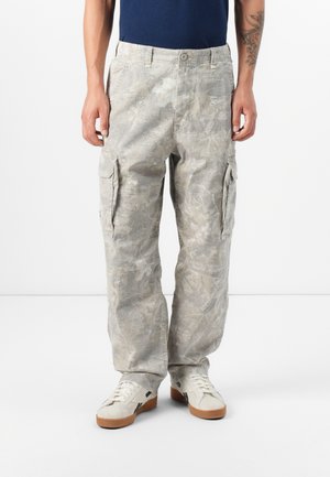 Mann trägt hellgraue Camouflage-Cargohosen mit Seitentaschen, weiße Sneakers und ein dunkelblaues Hemd, steht vor einem schlichten weißen Hintergrund.