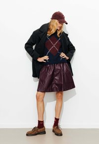 La tenue se compose d'un manteau noir, d'un pull en argyle bleu marine, d'une jupe en cuir bordeaux et de chaussures à imprimé léopard avec des chaussettes bordeaux.