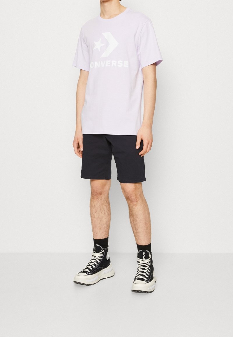 Personne portant un t-shirt Converse lavande clair, un short noir, des chaussettes noires et des baskets montantes noires Converse, debout devant un fond blanc.