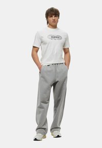 T-shirt bianco con grafica nera "GUESS", abbinato a pantaloni della tuta grigi. L'outfit presenta una vestibilità comoda e un tessuto morbido, indossato con sneakers bianche.