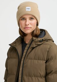 Brun pufferjakke med hette, med glidelås og teksturert stoff. Beige strikket lue med merkelapp, svart t-skjorte under.