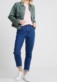 Chaqueta de denim verde sobre una camiseta rosa, combinada con jeans azules enrollados y zapatillas blancas. Incluye botones de metal y un ajuste relajado.