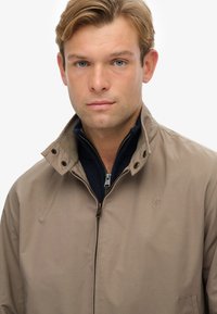 Beige jas met een hoge kraag, drukknoopsluiting en een rits aan de voorkant. Beschikt over een blauwe binnenlaag met een soepele textuur en subtiele logodetails.