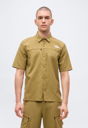 Άνδρας που φοράει ανοιχτό καφέ κοντομάνικο πουκάμισο με κουμπιά και ταιριαστό παντελόνι με τσέπες με φερμουάρ, στέκεται μπροστά σε απλό ανοιχτό φόντο.