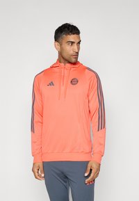 Lys oransje hettegenser med glidelås foran, hette og tre grå striper på hver erme. Har et rundt logo på brystet og ribbestrikkede mansjetter.