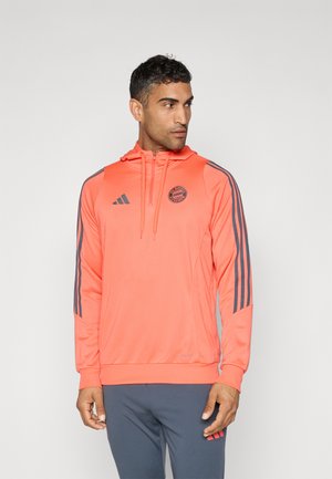Hoodie orange vif avec une fermeture éclair sur le devant, une capuche et trois rayures grises sur chaque manche. Présente un logo rond sur la poitrine et des poignets côtelés.