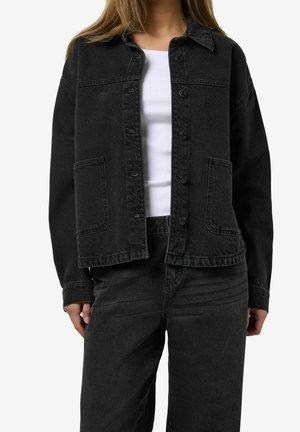 Vrouw draagt een zwarte spijkerjasje en bijpassende zwarte jeans met een wit geribd tanktop eronder, handen ontspannen langs het lichaam.