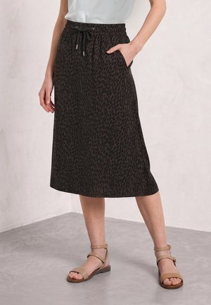 Femme portant une jupe noire mi-longue avec un subtil imprimé léopard, un haut blanc et des sandales beiges à bride cheville, debout sur un sol gris.
