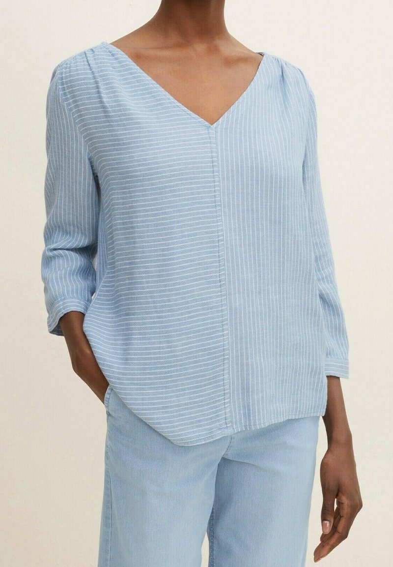 Femme portant un chemisier bleu clair à fines rayures avec un col en V et des manches trois-quarts, et un pantalon assorti bleu clair, debout avec une main dans la poche.