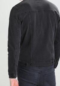 Osoba oblečená v přiléhavé černé džínové bundě a tmavých kalhotách, zobrazena zezadu na jednoduchém světle pozadí.