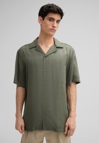 Chemise à manches courtes, en tissu léger olive. Présente un devant à boutons, un col et une poche poitrine unique. Texture douce.