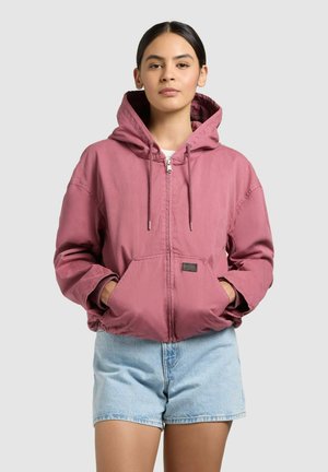 Mlada ženska z rokami v žepih, oblečena v jopico z zadrgo v prašno rožnati barvi s kapuco in svetlo modre džins kratke hlače na enobarvnem ozadju.