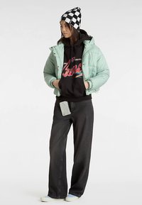 Vaaleanvihreä toppatakki, musta huppari värikkäällä tekstillä, mustat leveälahkeiset farkut ja ruudullinen pipo. Älypuhelin farkkujen taskussa.