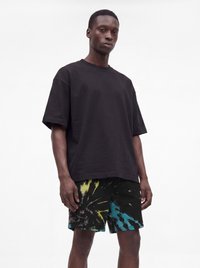 Ung mand iført en sort oversized t-shirt og sorte tie-dye shorts med gule og blå mønstre, stående mod en hvid baggrund.