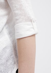 Bílá lněná blůza s vyhrnutými rukávy, se zajímavým detailním zapínáním. Materiál je lehký a mírně průhledný s texturovaným povrchem.