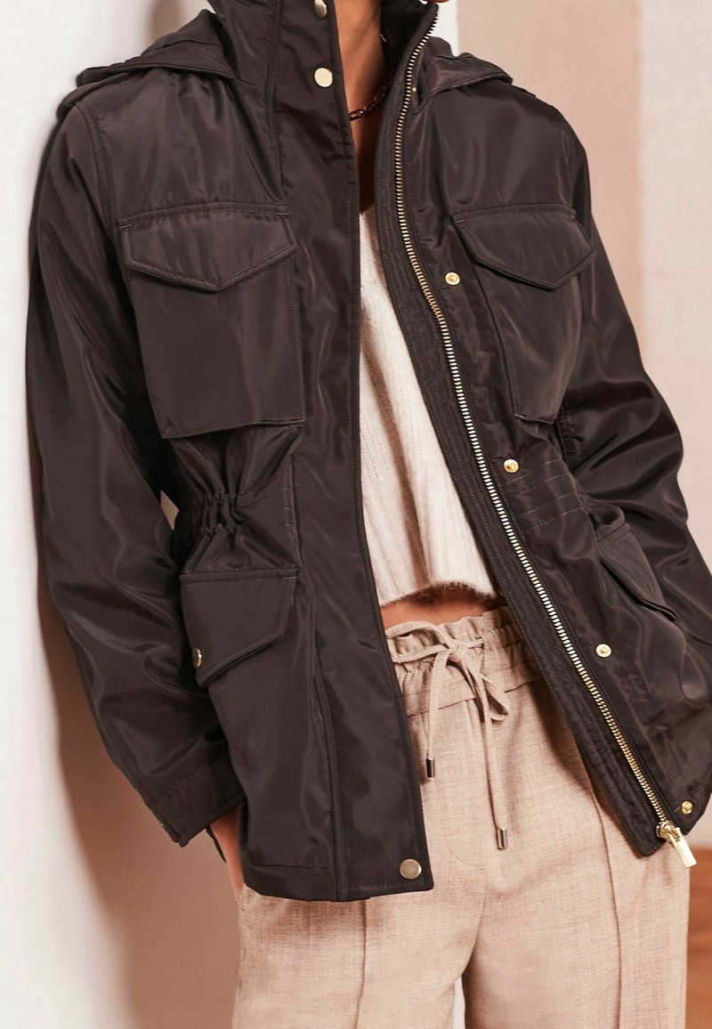 Veste noire zippée avec poches avant à rabat portée sur un pull court blanc et un pantalon beige avec cordon de serrage.