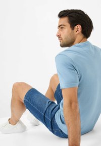 Lyseblå t-shirt med korte ærmer, parret med mørkeblå denimshorts og hvide sneakers. Model sidder, set fra siden og bagfra.