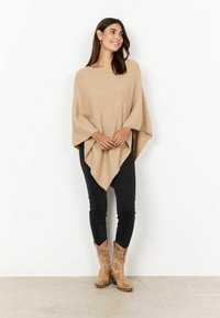 Poncho de culoare marron deschis cu textură strălucitoare, design cu falduri și formă triangulară, asortat cu pantaloni negri skinny și cizme de cowboy de culoare bej.