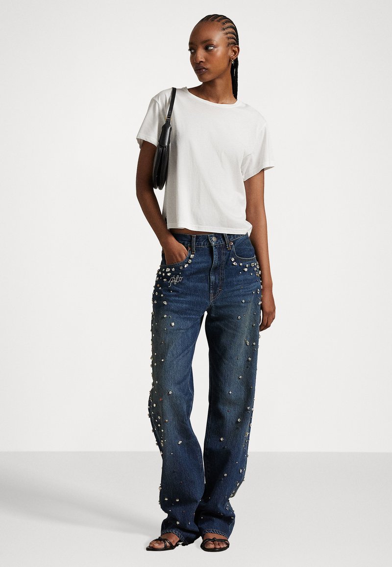 Weißes, gekürztes T-Shirt kombiniert mit hoch taillierten blauen Jeans, die mit verzierten Details und einem lässigen Schnitt ausgestattet sind. Eine schlichte schwarze Handtasche rundet den Look ab.