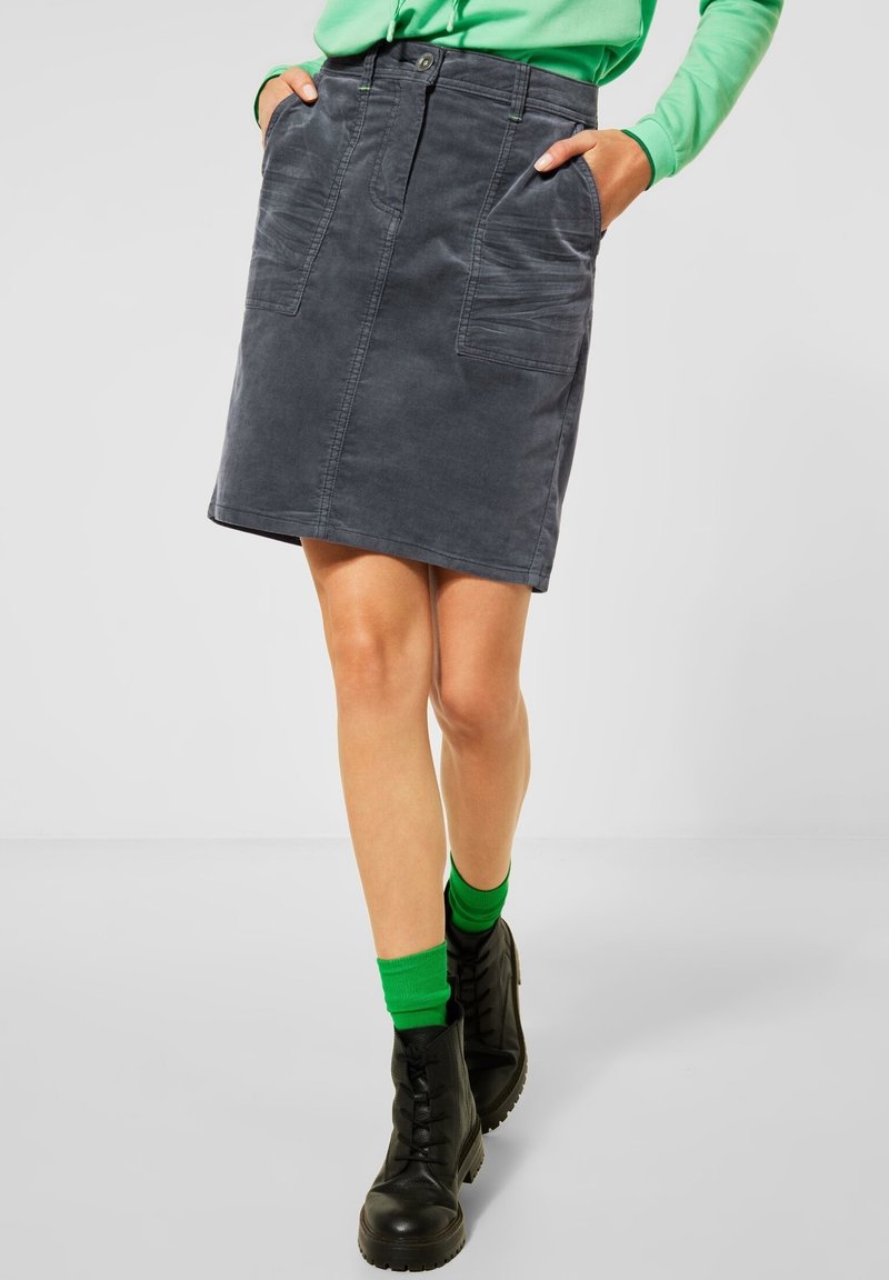 Jupe en denim gris avec deux poches avant, couture visible et fermeture à boutons. Portée avec des bottes noires et des chaussettes vertes.