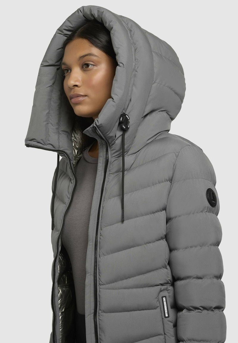 Eine Frau trägt eine graue wattierte Jacke mit einer großen Kapuze und einem schwarzen Reißverschluss über einem dunkelgrauen Oberteil. Sie steht mit dem Gesicht nach links vor einem schlichten Hintergrund.