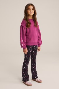 Violeta спортивная свитер ar puffed piedurknēm, pārī ar melnām legingām, kurām ir rozā debesu raksti ar mēness un zvaigznēm. Apavi ir gaiši rozā.