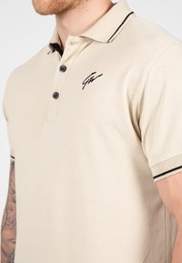 Beige poloshirt met een gestructureerd design, drie zwarte knopen en een zwart geborduurd logo op de linkerborst, met een kraag met contraststrepen.