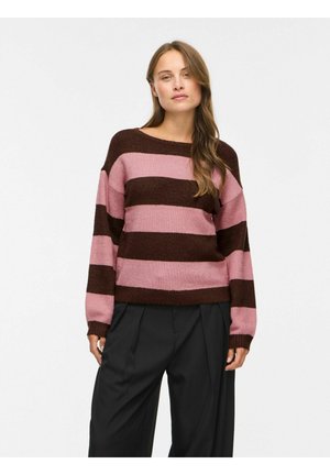 Femme portant un pull ample rayé rose et brun foncé à manches longues et un pantalon noir à jambes larges, debout devant un fond uni.