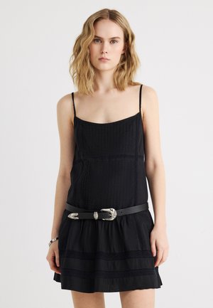 Młoda kobieta o falowanych blond włosach, ubrana w czarną sukienkę bez rękawów z cienkimi ramiączkami oraz czarny pasek z srebrną klamrą.