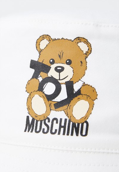 Gorra de tela blanca con un oso de dibujos animados que sostiene el texto "JUGUETE". El oso es marrón con acentos negros, sobre un fondo blanco liso.