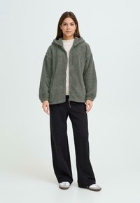 Grüne Fleecejacke mit Kapuze, Frontreißverschluss, Kordelzügen und gerippten Bündchen. Getragen über einem cremefarbenen Pullover und kombiniert mit schwarzen Hosen.