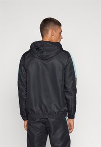Chaqueta atlética negra con capucha, puños elásticos y cordón ajustable. Presenta rayas laterales turquesas y una textura suave y brillante.