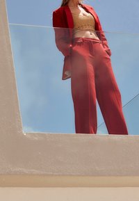 Rode op maat gemaakte blazer over bijpassende hoge taille broek, gecombineerd met een gestructureerde gouden crop top, tegen een heldere blauwe lucht.