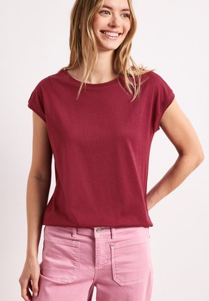 Smilende kvinde iført en burgunder kortærmet top og lyserøde jeans, stående med den ene hånd på hoften mod en ensfarvet baggrund.