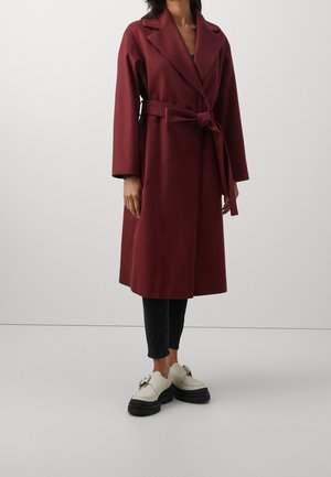 Femme portant un long manteau bordeaux ceinturé, un pantalon court noir et des mocassins blancs épais, debout devant un fond clair uni.